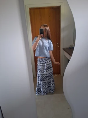 Långkjol med blåvitt mönster - Säljer en långkjol med snyggt blåvitt mönster. Kjolen har en flowy passform och är perfekt för dig som gillar boho-vibbar. Materialet känns lätt och skönt, perfekt för varmare dagar. Matcha med en croppad topp för en chill look.