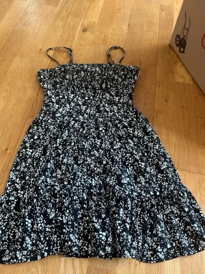 Blommig sommarklänning från NA-KD - Söt svart klänning med vita blommor från NA-KD x Pamela. Klänningen har smala axelband, smock upptill och en lätt utställd kjol. Perfekt för varma dagar och ger en fräsch och ungdomlig look.