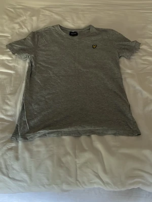 Grå t-shirt från Lyle & Scott - Säljer en stilren grå t-shirt från Lyle & Scott med klassisk rund hals och den ikoniska gula loggan broderad på bröstet. Tillverkad i mjuk bomull som känns skön mot huden. Perfekt till jeans eller shorts för en enkel och clean look.