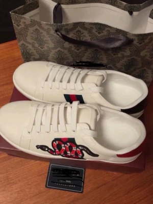 Vita sneakers med orm och röd detalj - Snygga vita sneakers med broderad orm på sidan och klassiska röd-blå ränder. Skorna har vita snören och en röd detalj på hälen. Låg modell med stilren design och coola detaljer som sticker ut.