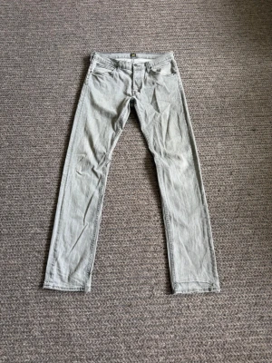 Lee jeans - Storlek 32/34. Säljer ett par ljusgrå jeans med rak passform och klassisk femficksdesign. Jeansen har normal midja och dragkedjegylf. Perfekta för en clean och stilren look.