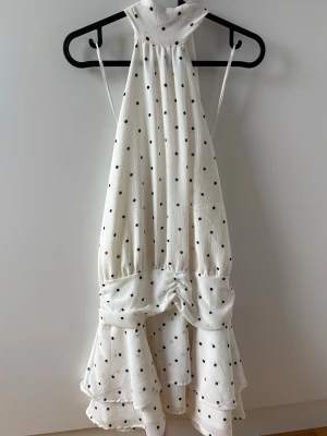 Vit prickig volangklänning - Supersöt vit klänning med svarta prickar och volangdetaljer. Klänningen har halterneck, öppen rygg och omlottdesign baktill. Perfekt för sommarens alla tillfällen och har en luftig, flowy passform.