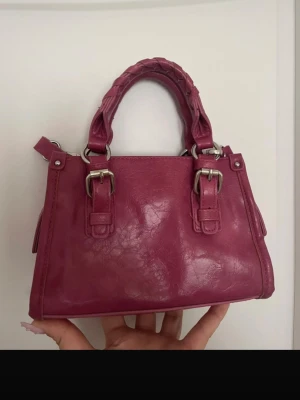 Rosa handväska med flätat handtag - Snygg rosa handväska i glansigt konstläder med två flätade handtag och silverfärgade metalldetaljer. Väskan har en boxig form och dekorativa spännen framtill. Perfekt accessoar för att lyfta din outfit.
