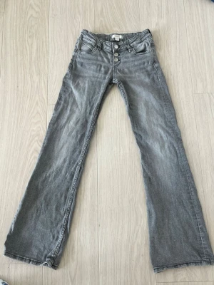 Grå bootcut jeans med knappar - Snygga grå lågmidjade, bootcut jeans med tre synliga knappar i midjan och klassiska fem fickor. Jeansen har en lätt utsvängd passform nedtill och är tillverkade i ett mjukt denimtyg. Är stora i storleken. Perfekta för en avslappnad och trendig look.
