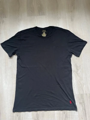 Polo Ralph Lauren T-shirt – Storlek L Slim Fit - Säljer en stilren och klassisk svart t-shirt från Polo Ralph Lauren i storlek Large (L). Modellen har en kroppsnära passform (Slim Fit) och är tillverkad i skön 100% bomull. Tröjan har den ikoniskt broderade röda hästloggan nertill i vänstra hörnet och en helt slät baksida. Tröjan är i fint begagnat skick utan skador eller missfärgningar. 