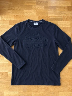 Svart långärmad tröja Superdry - Svart långärmad tröja från Superdry med rund hals och diskret broderad logga framtill. Tillverkad i mjukt bomullsmaterial som känns skönt mot huden. Enkel och stilren design som funkar till det mesta.