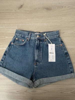 Högmidjade blå denimshorts - Snygga högmidjade denimshorts i mellanblå tvätt med uppvikta benslut och klassiska fem fickor. Perfekta för sommaren och har en tidlös look som passar till det mesta. Stängs med knapp och dragkedja framtill.