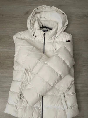 Vit pufferjacka från Tommy Hilfiger - Snygg vit pufferjacka från Tommy Hilfiger med huva och dragkedja framtill. Jackan har två sidofickor med dragkedja och diskret logga på bröstet. Perfekt för kalla vinterdagar och riktigt trendig look.