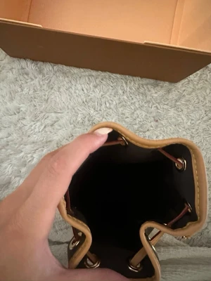 monogram bag - Stilren bucket handväska i brunt och beige med monogrammönster. Väskan har snörning upptill, guldfärgade metalldetaljer och detaljer i ljust skinn. Perfekt för dig som gillar trendiga accessoarer med lyxig känsla.
