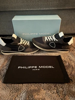 Philippe Model mörkblåa sneakers - Snygga mörkblåa och grå sneakers från Philippe Model med vita detaljer och vita skosnören. Skorna har en låg siluett, klassisk Philippe Model-logga på sidan och en mix av mocka och textilmaterial. Perfekta för en trendig och avslappnad stil.