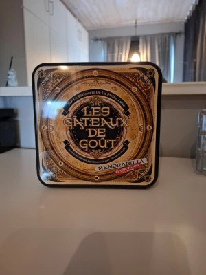 Les Gateaux De Gout memorabilia box - Säljer en fyrkantig samlarlåda med texten 'Les Gateaux De Gout' och 'Memorabilia Dark Moon'. Lådan är i metall med guldfärgad och svart design, och har detaljerade mönster på framsidan. Perfekt för dig som gillar unika samlarprylar eller K-pop merchandise.