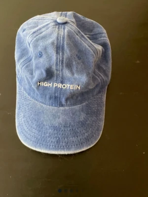 Blå keps med brodyr High Protein - Snygg blå keps från Star Nutrition med broderad text 'HIGH PROTEIN' framtill och 'WHEY-80' på sidan. Klassisk böjd skärm och justerbar passform. Perfekt accessoar för gymmet eller vardagslooken.