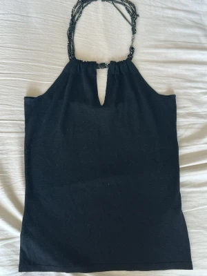 Svart halterneck topp med pärlband - Snygg svart halterneck topp med unika pärlband som går runt halsen. Toppen har en liten v-ringning framtill och är ärmlös med en tight passform. Perfekt för dig som vill ha något stilrent men ändå speciellt.