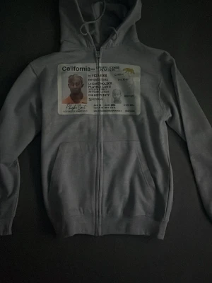 Grå hoodie med tryck - Grå zip-hoodie med stor tryckt bild av ett amerikanskt körkort på bröstet. Hoodien har huva med dragsko och två fickor framtill. Perfekt för dig som vill sticka ut med en unik och rolig design.