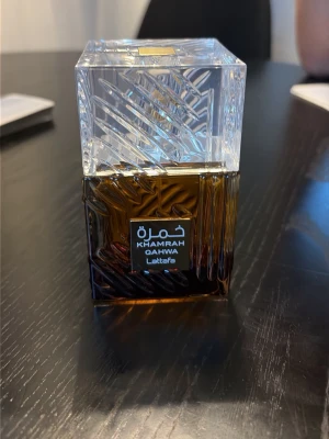 Lattafa Khamrah Qahwa parfym 100ml - Lattafa Khamrah Qahwa är en exklusiv parfym i en elegant fyrkantig glasflaska med mönstrad, transparent kork. Flaskan har en djup bärnstensfärg och levereras i en lyxig brun förpackning. Doften är Eau de Parfum och flaskan rymmer 100 ml.