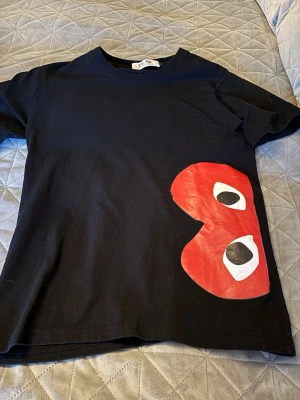 Svart Comme des Garçons PLAY t-shirt - Säljer en svart t-shirt från Comme des Garçons PLAY med det ikoniska röda hjärtat med ögon tryckt på sidan. T-shirten har rund hals och korta ärmar, perfekt för en clean och cool look.