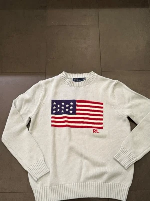Ljusgrå stickad tröja Ralph Lauren - Ljusgrå stickad tröja  Ralph Lauren med amerikansk flagga i blått, rött och vitt på bröstet samt RL-broderi. Tröjan har rund halsringning, ribbade muddar och nederkant. Perfekt för dig som gillar klassisk och stilren design.
