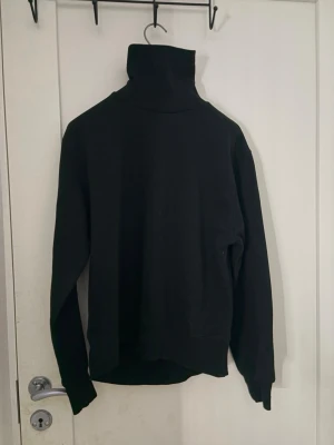 Svart polotröja med hög krage - En stilren svart polotröja med lång ärm och extra hög krage. Tröjan har en rak passform och är tillverkad i ett mjukt material som känns skönt mot huden. Perfekt för lager på lager och enkel att matcha med olika outfits.