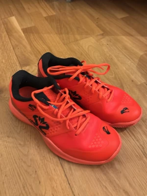 Röda padelskor med svart detalj - Säljer ett par snygga röda padelskor med svarta detaljer och svensk flagga på plösen. Skorna har snörning, låg profil och är tillverkade i syntetmaterial med ventilerande meshpartier. Perfekta för inomhussport eller gym.