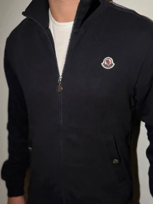  Moncler fullzip  - Snygg svart ziptröja från Moncler med logga på bröstet och coola röda, vita och svarta ränder längs ärmarna. Tröjan har hög krage, två fickor med knappar och ribbade muddar. Perfekt för en sportig och stilren look.