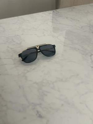Svarta solglasögon med gulddetaljer - Snygga solglasögon med svart båge och mörka glas. Ramen har coola gulddetaljer och en markerad brygga som ger en modern och trendig look. Perfekta för dig som vill sticka ut och addera lite edge till din stil.