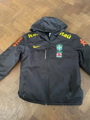 Svart Brasil Nike jacka med huva - Snygg svart jacka från Nike med Brasiliens landslagslogga och sponsorer i gult och orange. Jackan har huva, dragkedja framtill och två sidofickor med dragkedja. Perfekt för dig som gillar sportig stil och vill visa stöd för Brasilien.  Storlek XL men är slim fit och passar även till M. Ingen storleks lapp på den men storleken är xl 