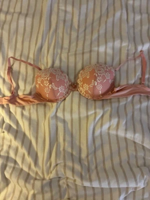 Rosa push-up bh med spetsdetaljer - Söt rosa push-up bh med vadderade kupor och vita spetsdetaljer i blommönster. Justerbara axelband och klassisk knäppning bak. Materialet är en kombination av mjuk textil och spets som ger en romantisk känsla.