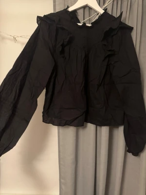 Svart blus med volangdetaljer - Snygg svart blus med volangdetaljer vid axlar och halsringning. Blusen har långa ärmar med resår vid ärmslut och en lös, luftig passform. Perfekt att styla med jeans eller kjol för en trendig look.