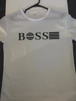 Vit t-shirt med BOSS-tryck - Snygg vit t-shirt med svart BOSS-tryck på bröstet. T-shirten har rund hals och korta ärmar. Materialet ser ut att vara mjukt och lätt, perfekt för en clean och stilren look.