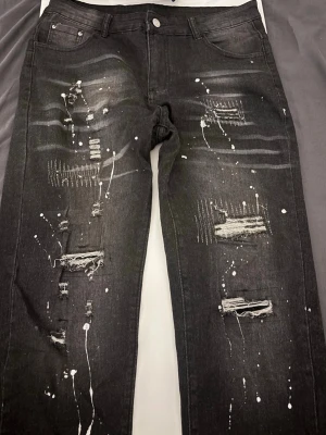Svarta distressed jeans med färgstänk - Säljer ett par svarta jeans med coola slitningar och vita färgstänk för en edgy look. Jeansen har fem fickor, normal midja och raka ben. Materialet är klassisk denim med en trendig, sliten finish och detaljer som ger en unik streetstyle-vibe.