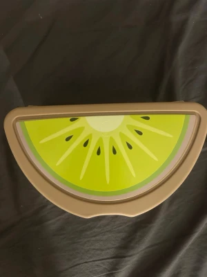 Matlåda i plast med kiwi-motiv - Säljer en halvcirkelformad matlåda i beige plast med ett färgglatt kiwi-motiv på locket. Insidan har ett separat fack för bestick och en medföljande spork. Perfekt för att ta med lunch eller snacks till skolan eller jobbet.