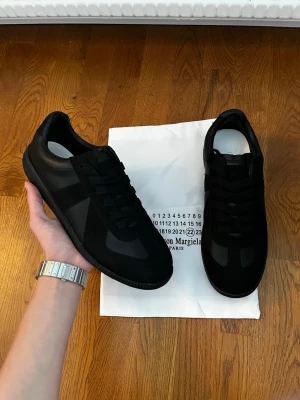 Svarta sneakers Maison Margiela - Snygga helsvarta sneakers från Maison Margiela i en mix av mocka och läder. Klassisk låg modell med svarta skosnören och diskret design. Perfekta för dig som gillar stilrena och tidlösa skor.