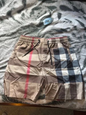 Beige-rutiga shorts med snörning - Säljer ett par beige shorts med klassiskt rutigt mönster i svart, vitt, brunt och rött. Shortsen har elastisk midja med svart snörning och sidofickor samt en bakficka. Perfekta för sommaren och riktigt snygga till streetwear-stilen. Passar M och L