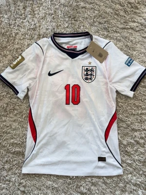 England Bellingham matchtröja Nike - Säljer en vit England fotbollströja från Nike med nummer 10 och Bellingham på ryggen. Tröjan har svarta och röda detaljer, broderat landslagsemblem och korta ärmar. Perfekt för dig som vill stötta England på match eller träning.