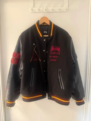Svart Stussy bomberjacka med brodyr - Säljer en svart bomberjacka från Stussy med broderade detaljer och patchar. Jackan har läderärmar, ribbade muddar i svart, gult och rött samt brodyr på bröstet och ryggen. Fickor framtill och coola detaljer som 'International Stussy Tribe' och siffror på ärmarna. Vid snabb affär kan pris diskuteras. 