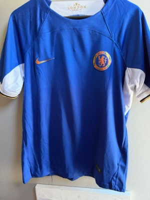 Chelsea FC matchtröja Mudryk #10 - Säljer en blå Chelsea FC matchtröja från Nike med Mudryk och nummer 10 på ryggen. Tröjan har klubbmärke och Nike-logga framtill, samt Premier League-märke på ärmen. Tillverkad i lätt och ventilerande material, perfekt för fotboll eller träning.
