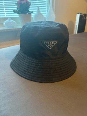 Prada bucket hat - Snygg svart bucket hat från Prada med det klassiska trekantiga metallmärket framtill. Hatten är tillverkad i ett glansigt nylonmaterial som ger en modern och clean look. Perfekt accessoar för att lyfta din outfit och ge en exklusiv touch.