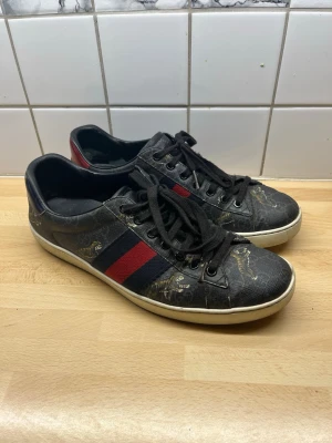 Svarta gucci ace sneakers  - Snygga svarta gucci ace sneakers med klassisk röd och blå rand på sidan. Skorna har svarta skosnören, vit sula och detaljer i guld med tigrar på ovandelen. Materialet ser ut som konstläder med tryckt mönster. Perfekta för dig som vill sticka ut lite extra. Det är bara att höra av sig vid frågor och funderingar!