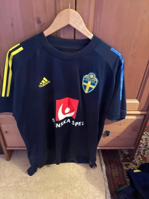 Svenska landslagets fotbollströja - Mörkblå fotbollströja med gula och blå detaljer, Svenska Spel-logga och svenska landslagets emblem på bröstet. Adidas-logga på framsidan och klassiska ränder på axlarna. Perfekt för fotbollsträning eller att heja på Sverige.