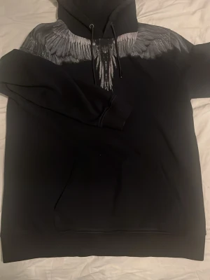 Marcelo Burlon Hoodie - Säljer en svart hoodie med stor huva och snyggt print av gråa vingar över axlarna, både fram och bak. Hoodien har dragsko i huvan och en klassisk magficka. Perfekt för dig som vill sticka ut med en unik design.