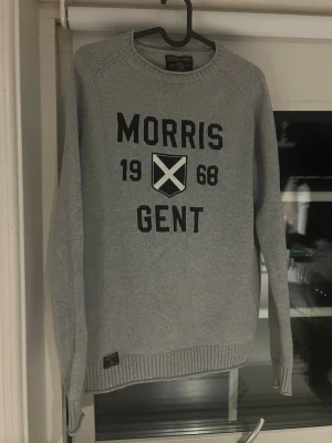Grå stickad tröja från Morris - Snygg grå stickad tröja från Morris med texten 'Morris Gent 1968' och ett tryckt emblem på bröstet. Tröjan har rund halsringning, långa ärmar och ribbade muddar vid ärmslut och nederkant. Perfekt för en stilren vardagslook. Kan skicka flera bilder