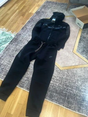 Svart Nike loungewear set - Snyggt svart loungewear set från Nike med zip hoodie och matchande byxor. Setet har diskreta Nike-loggor, dragsko i midjan och muddar vid benslut. Perfekt för chill dagar hemma eller när du vill vara bekväm men ändå se fräsch ut.