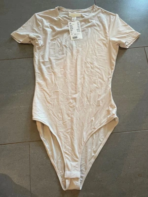 Beige kortärmad body XS - En stilren beige body med korta ärmar och rund hals. Tillverkad i mjukt och stretchigt material som sitter skönt mot kroppen. Perfekt att styla med jeans eller kjol för en enkel och snygg look.