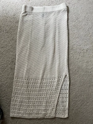 Beige strandkjol - Snygg beige stickad långkjol med dekorativt hålmönster nedtill och en liten slits på sidan. Kjolen har en ribbad midja och är perfekt för dig som gillar en avslappnad men trendig stil.