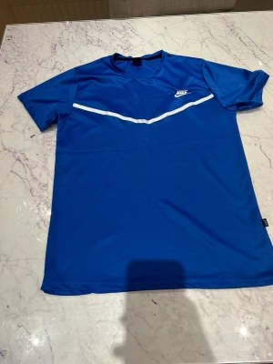 Blå Nike t-shirt med vit detalj - Säljer en blå t-shirt från Nike med en vit, böjd linje över bröstet och Nike-loggan på vänster sida. T-shirten har korta ärmar och är gjord i ett lätt och sportigt material som passar perfekt för aktiva dagar.