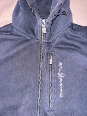 Marinblå hoodie från Sail Racing - Säljer en marinblå hoodie från Sail Racing med dragkedja framtill och en extra bröstficka med zip. Hoodien har justerbar huva med snören och Sail Racing-logga tryckt på bröstet. Mjuk insida och sportig look, perfekt för chill dagar.