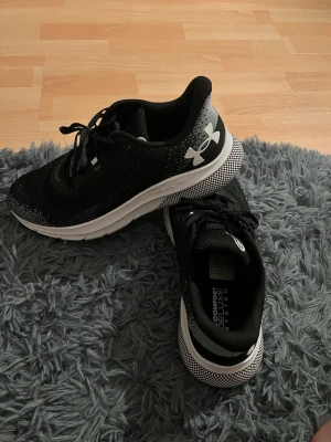 Svarta sneakers från Under Armour - Snygga svarta sneakers från Under Armour med vit sula och logga på sidan. Ovandelen är i meshmaterial för bra andningsförmåga och komfort. Skorna har klassisk snörning och en tjock, stötdämpande sula som passar perfekt till en aktiv livsstil.