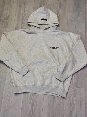  Essentials  - Snygg ljusgrå hoodie från Essentials Fear of God med svart logga på bröstet och stor text på ryggen. Klassisk passform med huva och ribbade muddar. Perfekt för en avslappnad och trendig streetwear-look.