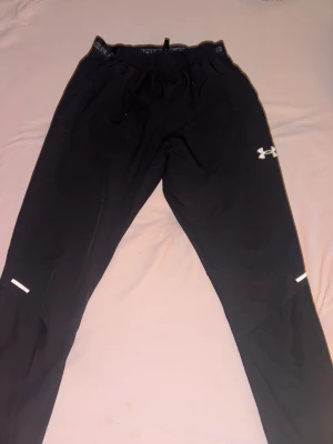 Svarta träningsbyxor Under Armour - Säljer ett par svarta träningsbyxor från Under Armour med resår och dragsko i midjan. Byxorna har vita logodetaljer och reflexdetaljer på benen. Perfekta för löpning eller gym. Tillverkade i ett lätt och stretchigt material.