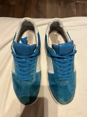Blå och vita valentino skor - Snygga sneakers i blå mocka med vita detaljer och vit sula. Skorna har en unik kamouflagemönstrad undersida och silverfärgade nitar runt sulan. Perfekta för dig som vill sticka ut med din stil.